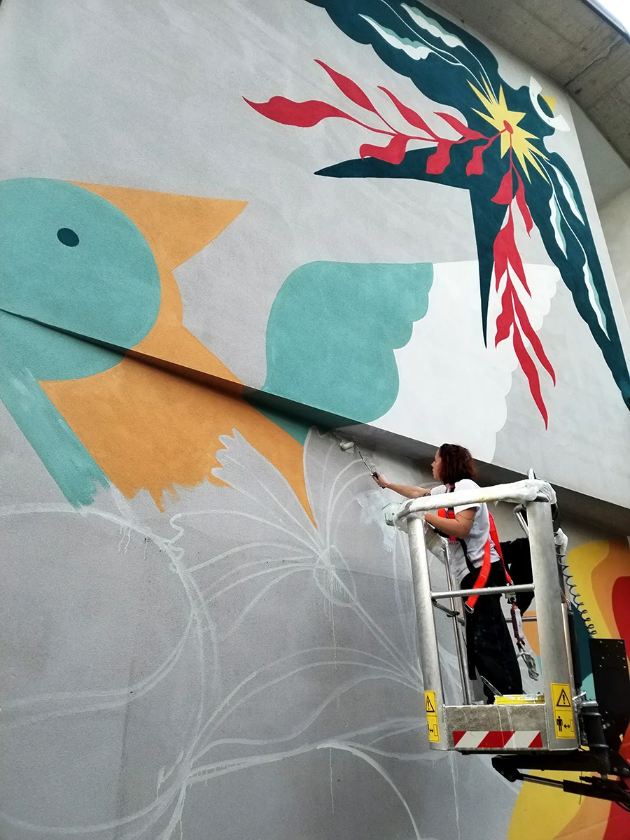 Dettaglio del murale 'Ad ali spiegate' di Rosmunda a Rovigo: l'artista al lavoro su una piattaforma elevatrice per rifinire i dettagli di una figura zoomorfa dalle forme ampie e colori vivaci.