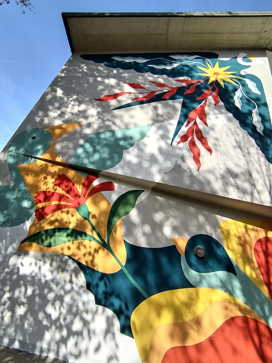 Altra foto con visuale dal basso dell'opera Ad Ali spiegate. L'ombra degli alberi di fronte si staglia sul murale.