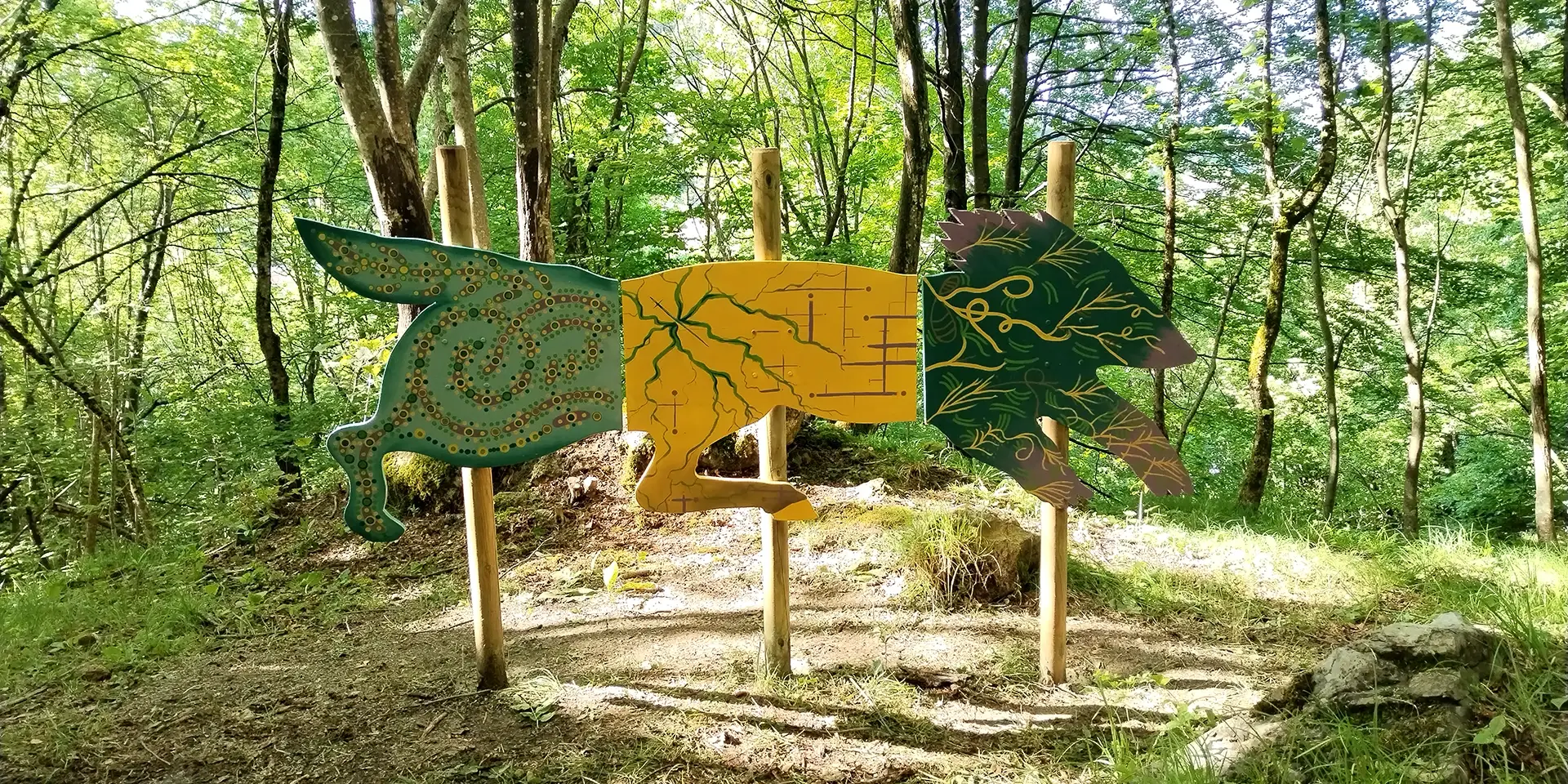 Veduta d'insieme della scultura in legno sagomata a forma di Chimera installata in un bosco, con il corpo suddiviso in tre pannelli decorati con motivi organici e geometrici nei toni del giallo oro, verde e ocra, sorretti da pali di legno tra gli alberi.