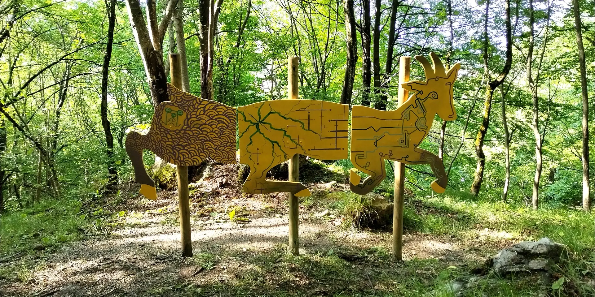 Veduta d'insieme della scultura in legno sagomata a forma di Chimera installata in un bosco, con il corpo suddiviso in tre pannelli decorati con motivi organici e geometrici nei toni del giallo oro, verde e ocra, sorretti da tre pali di legno tra gli alberi.