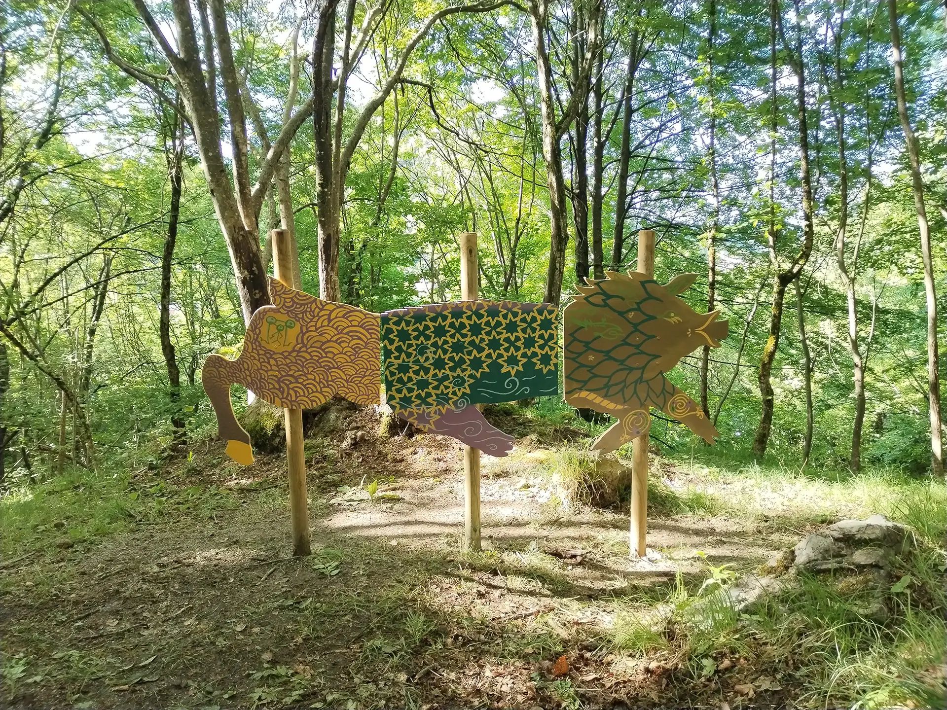 Veduta d'insieme della scultura in legno sagomata a forma di Chimera installata in un bosco, con il corpo suddiviso in tre pannelli decorati con motivi organici e geometrici nei toni del giallo oro, verde e ocra, sorretti da tre pali di legno tra gli alberi.
