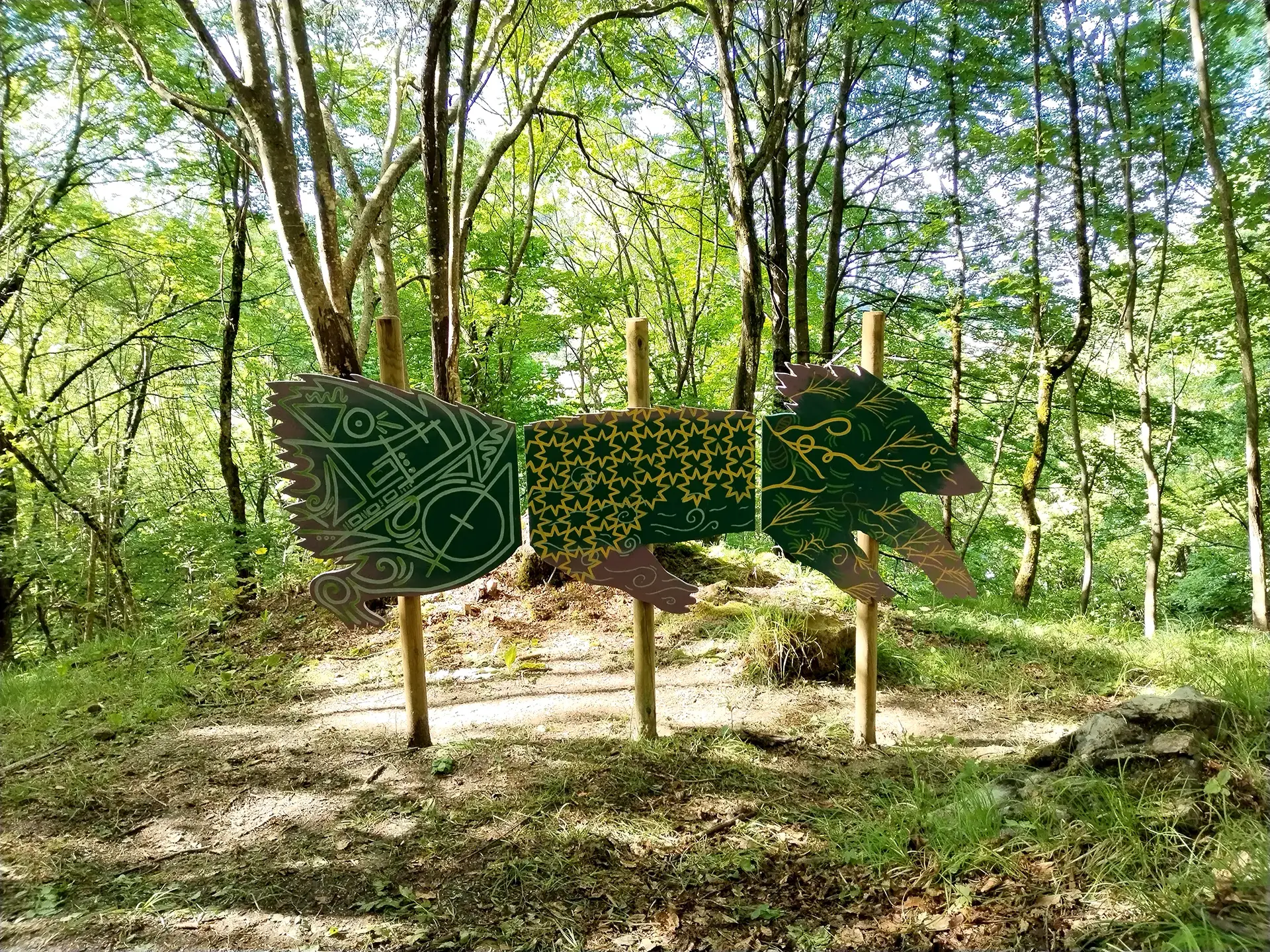 Veduta d'insieme della scultura in legno sagomata a forma di Chimera installata in un bosco, con il corpo suddiviso in tre pannelli decorati con intricati motivi geometrici e organici di colore verde scuro e bianco su sfondo ocra, sorretti da tre pali di legno tra la fitta vegetazione.