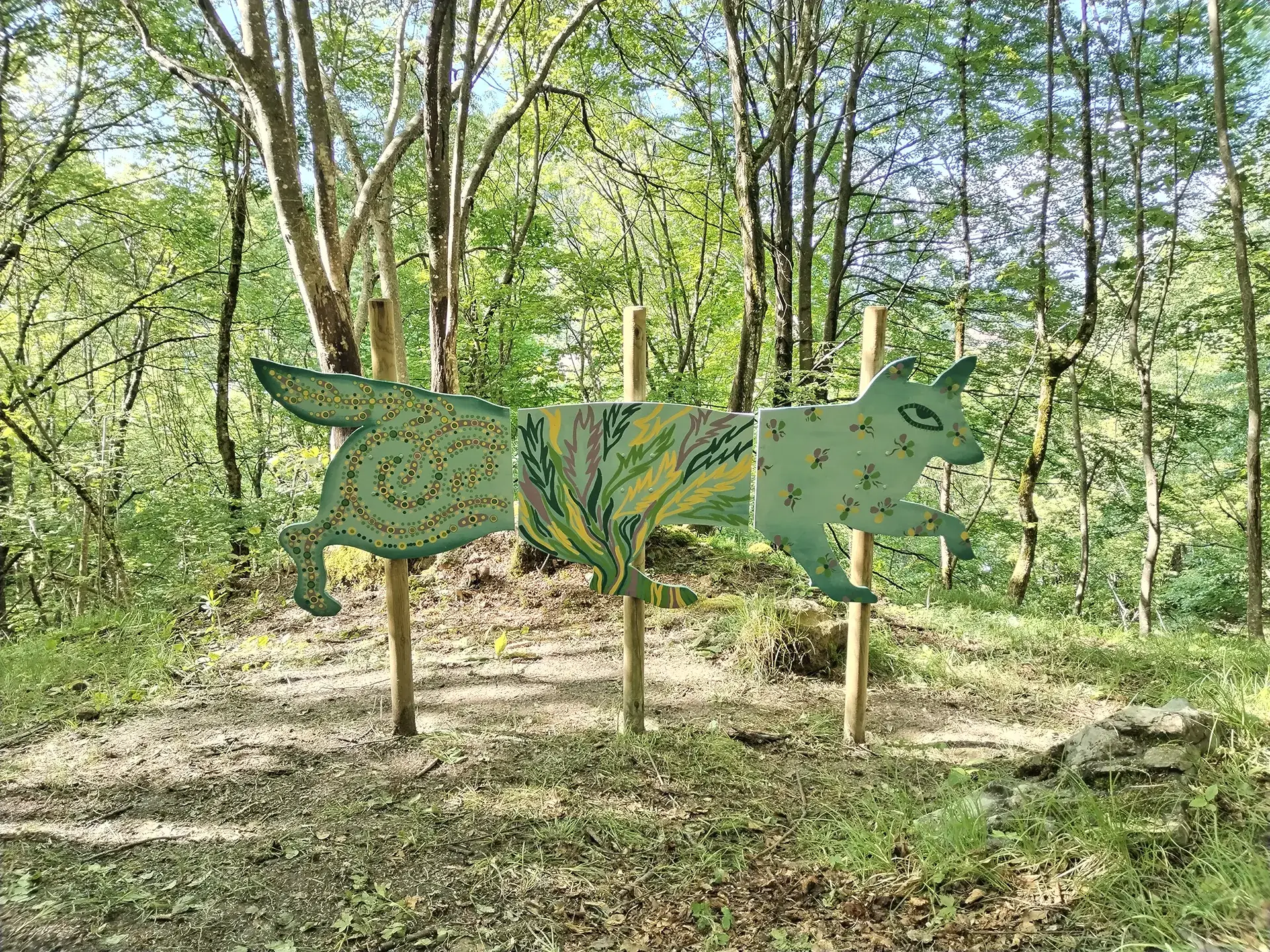 Installazione artistica nel bosco che raffigura una creatura fantastica stilizzata, la Chimera, composta da tre pannelli in legno sagomati e decorati con motivi organici floreali nei toni del verde chiaro e scuro su sfondo grigio-azzurro, sorretti da tre pali verticali tra gli alberi.