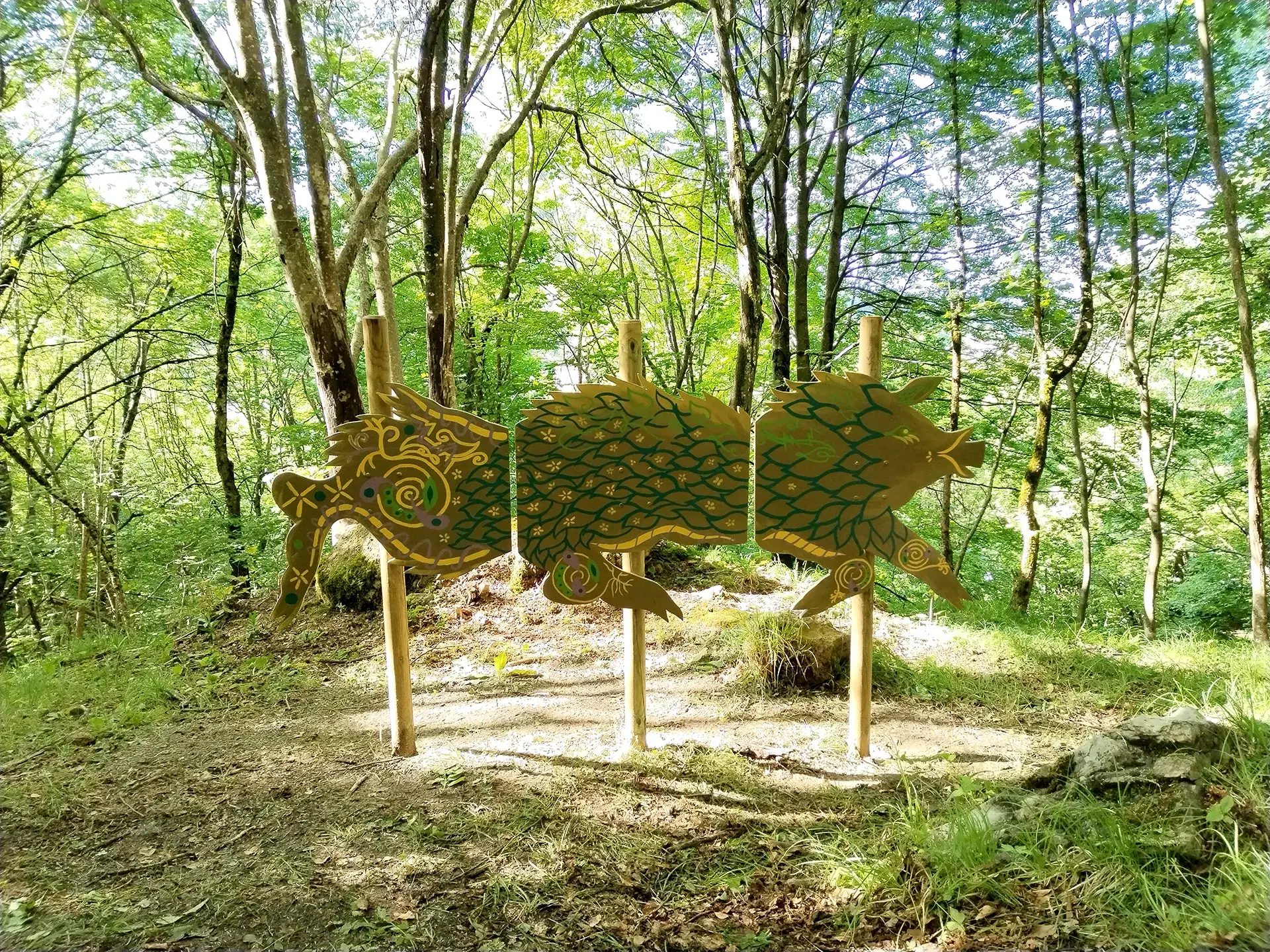 Installazione artistica nel bosco che raffigura una creatura fantastica stilizzata, la Chimera, composta da tre pannelli in legno sagomati e decorati con motivi organici e squame nei toni del verde e dell'oro, sorretti da tre pali verticali tra gli alberi.