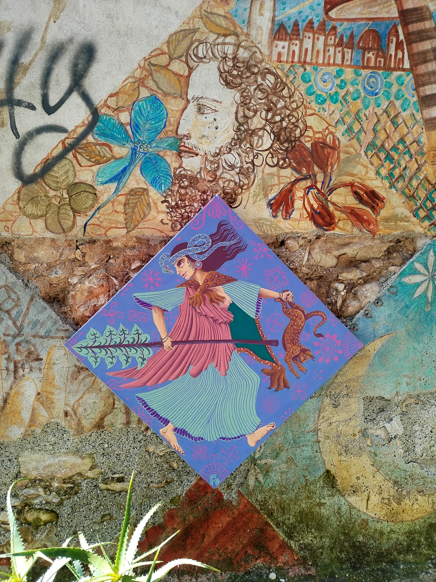 Opera d'arte contemporanea di Rosmunda intitolata "Ekstasis", raffigurante una figura femminile mitologica su una tela romboidale azzurra posizionata su una parete texturizzata con motivi floreali. La scena presenta una divinità in movimento con vesti fluide e un bastone rituale, circondata da decorazioni organiche e tonalità terrose.