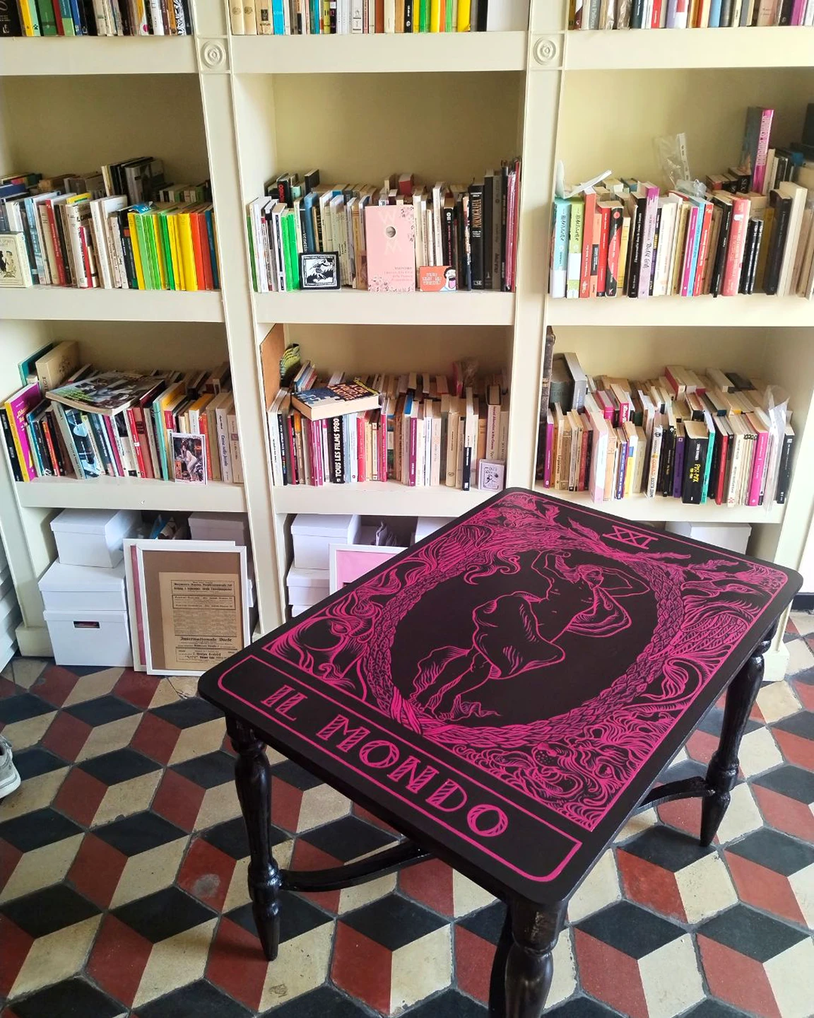 Veduta d'insieme del tavolo "Il mondo queer" di Rosmunda all'interno del Centro Aldo Mieli; il piano in legno, decorato con smalto fucsia su fondo nero, è circondato da sedie e posizionato davanti a una libreria a parete colma di volumi e materiale documentario.