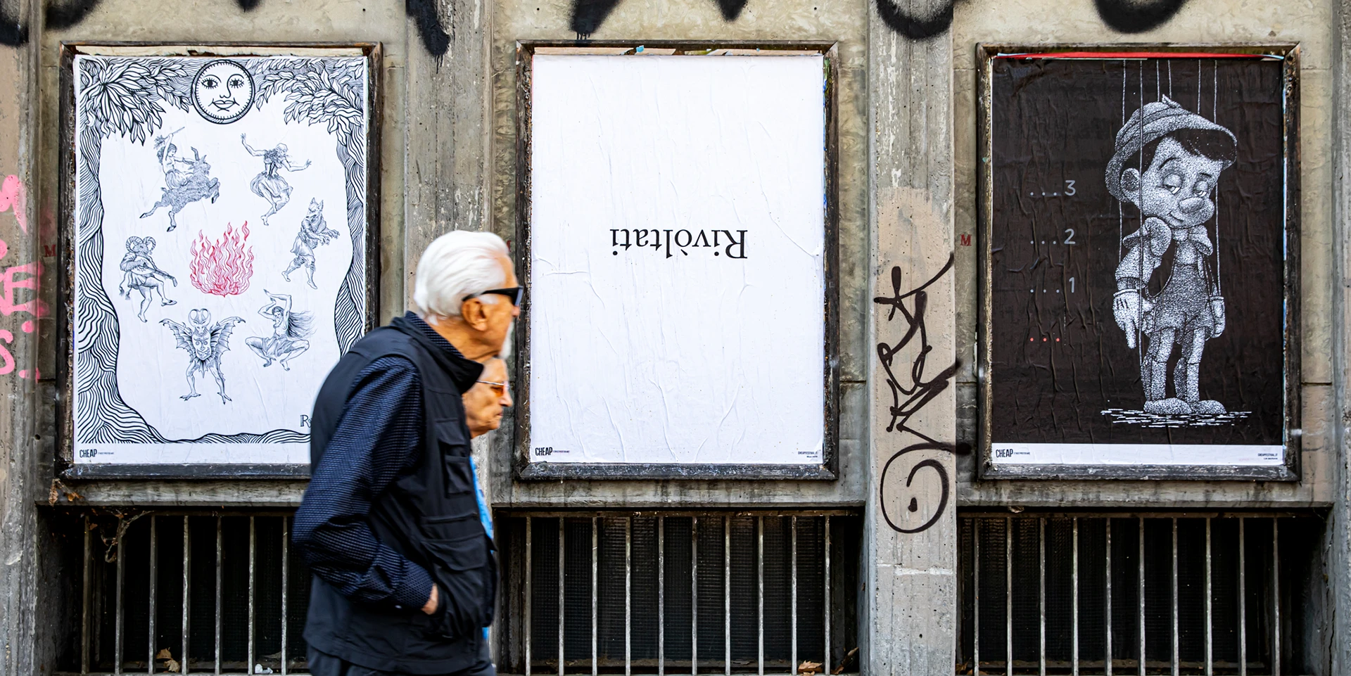 Fotografia di un intervento di street poster art a Bologna per il CHEAP Festival. Tre manifesti sono affissi su una parete urbana grigia: a sinistra l'opera "Il Sabba" di Rosmunda in bianco e nero con un fuoco rosso centrale, al centro un manifesto con la scritta sottosopra "Rivoltati", e a destra un'illustrazione di un Pinocchio malinconico. Due passanti camminano davanti alle opere.