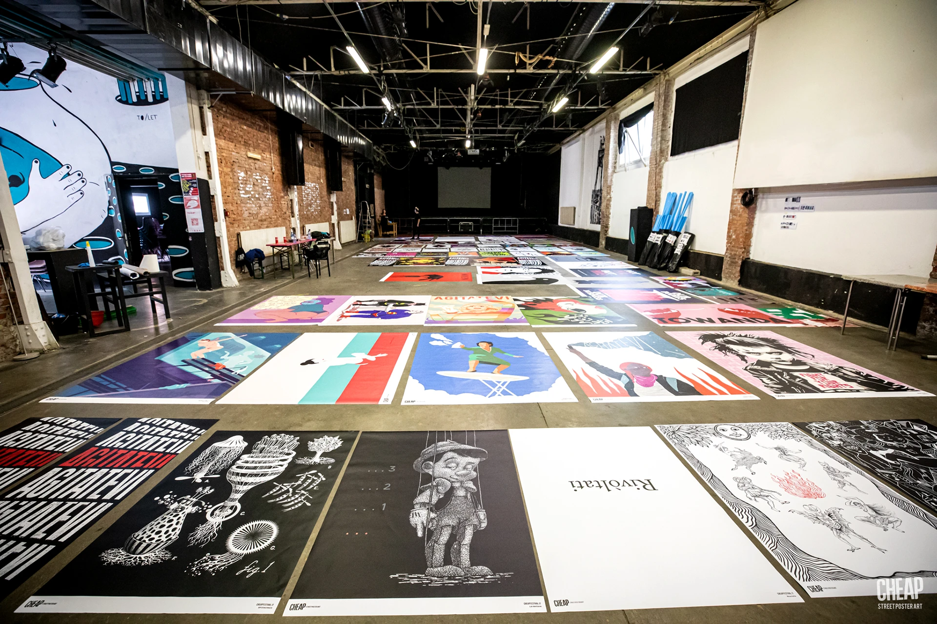 Fotografia scattata all'interno di un ampio spazio industriale (DUMBO Bologna) durante la fase di preparazione del CHEAP Festival. Numerosi manifesti colorati sono stesi a terra pronti per l'affissione; in primo piano si vedono le opere di Rosmunda in bianco e nero.