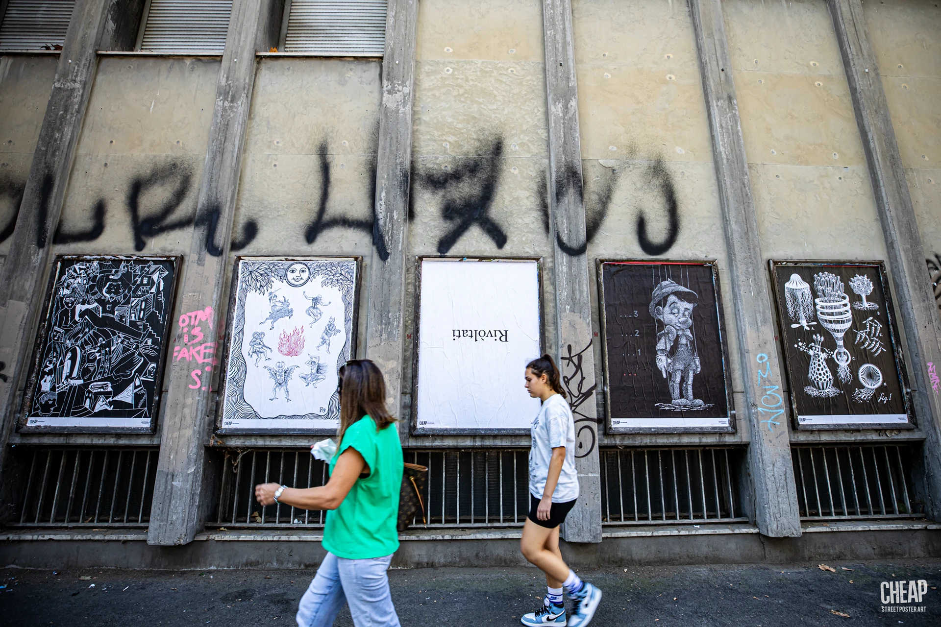 Veduta più ampia dell'affissione CHEAP a Bologna. Una serie di cinque poster artistici decora una bacheca pubblica sotto un muro caratterizzato da graffiti. Due donne camminano sul marciapiede adiacente, evidenziando l'integrazione dell'opera di Rosmunda nel tessuto quotidiano della città.