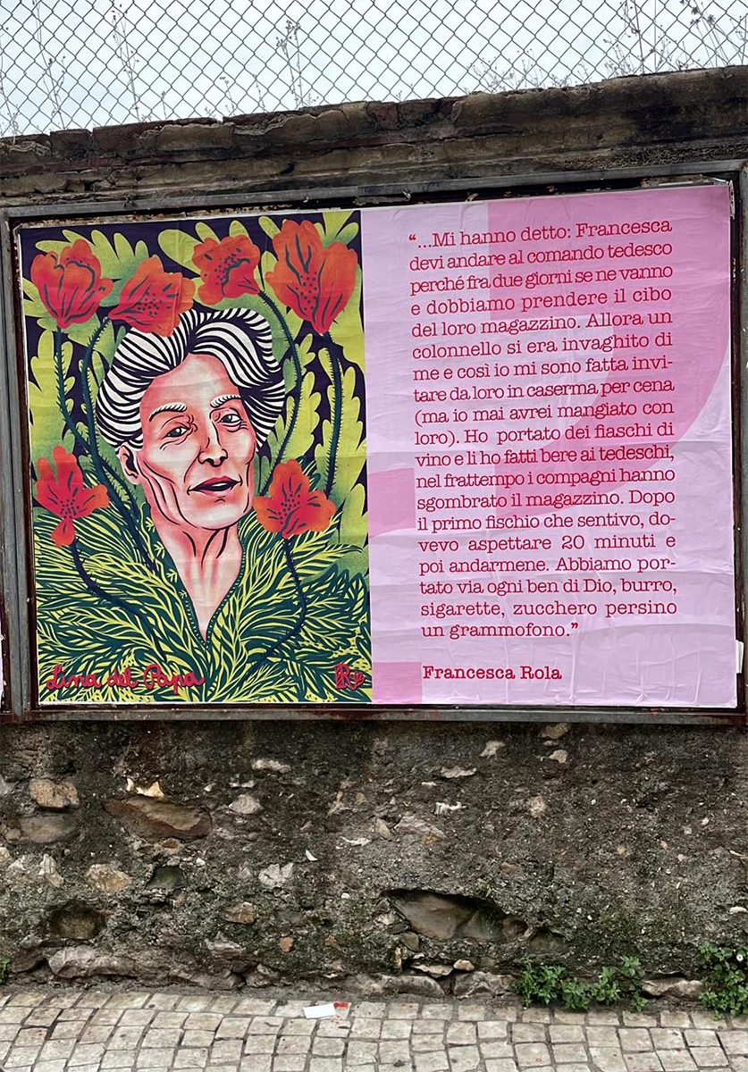 Fotografia di un'affissione pubblica su un muro di pietra a Carrara. Il poster di Rosmunda ritrae Lina Del Papa circondata da fiori rossi e foglie verdi; accanto, un manifesto rosa riporta una testimonianza storica del 7 luglio 1944. L'opera si integra nel paesaggio cittadino come un atto di memoria viva.