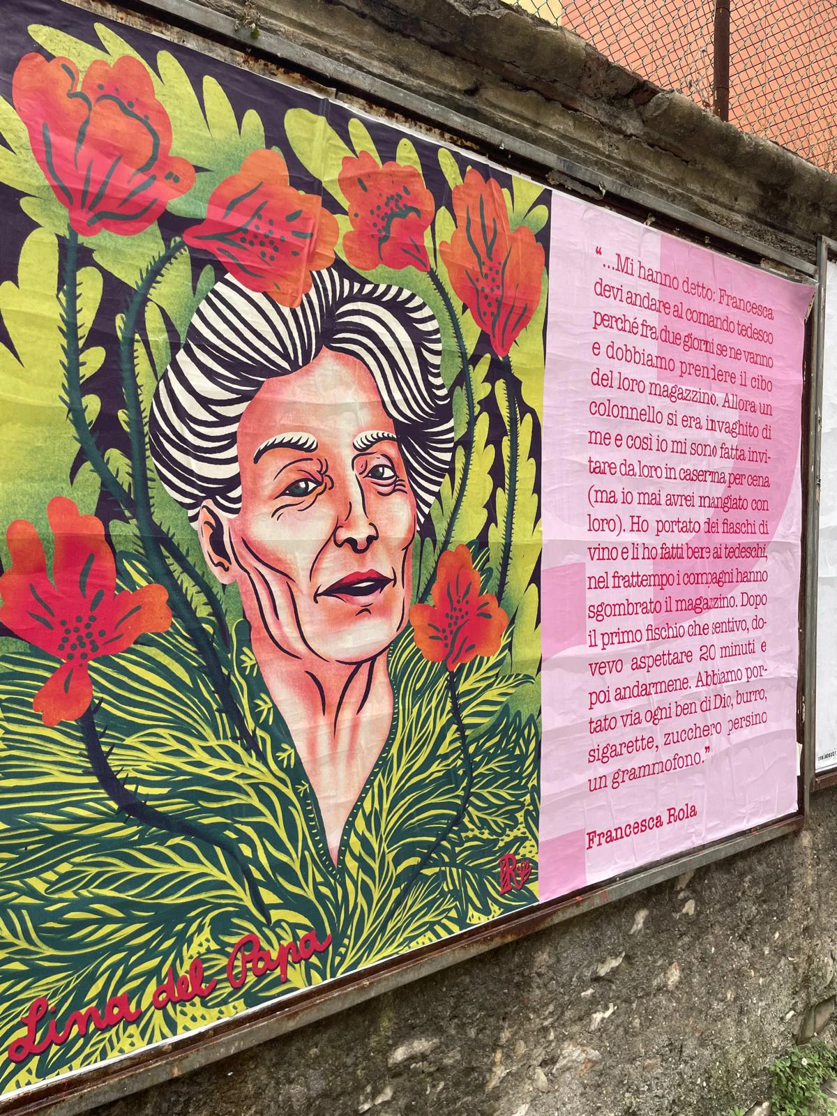 Veduta ravvicinata del poster dedicato a Lina Del Papa affisso all'aperto. La luce del giorno mette in risalto i colori accesi dell'illustrazione — il rosso dei fiori e il verde della vegetazione — che contrastano con la superficie materica e irregolare della parete urbana sottostante.