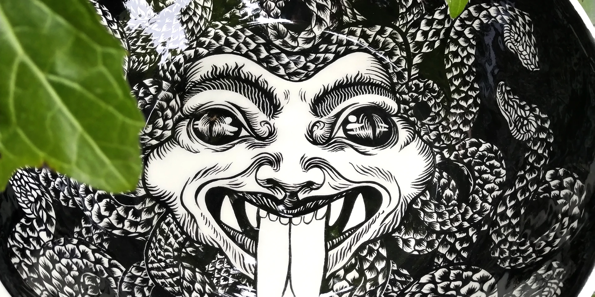 Dettaglio dell'opera in ceramica "Medusa" di Rosmunda che mostra in primo piano il volto della Gorgone dipinto all'interno di una ciotola. L'illustrazione in bianco e nero mette in risalto lo sguardo ipnotico, i denti aguzzi con la lingua biforcuta sporgente e la complessa trama di serpenti squamati che si intrecciano fittamente intorno al capo.