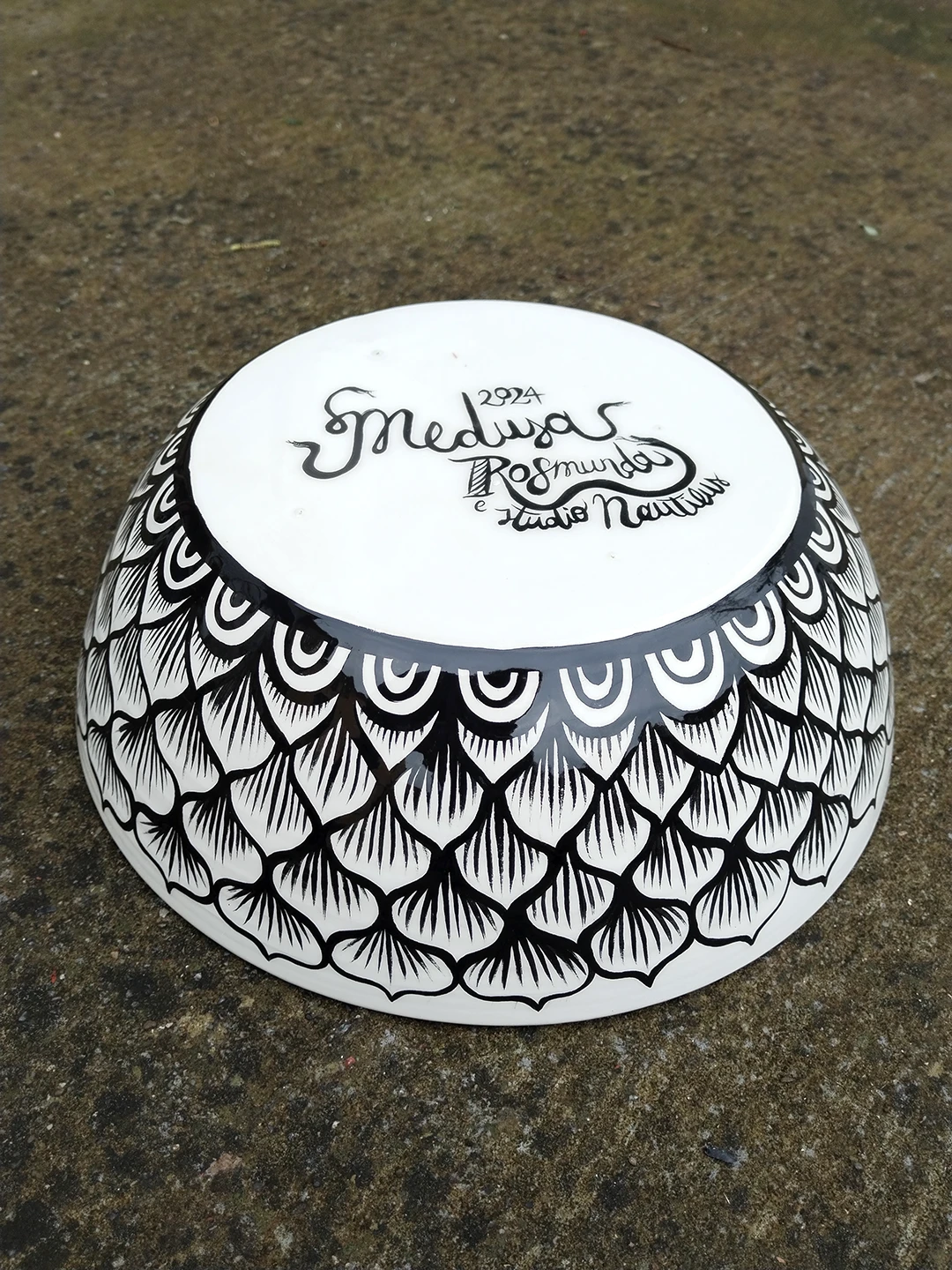 Veduta della base esterna della ciotola in ceramica "Medusa". La superficie è decorata con un motivo a squame dipinto a mano, mentre sul fondo bianco sono riportati il titolo dell'opera, la firma dell'artista Rosmunda e il nome dello studio Nautilus con cui è stata realizzata la collaborazione.