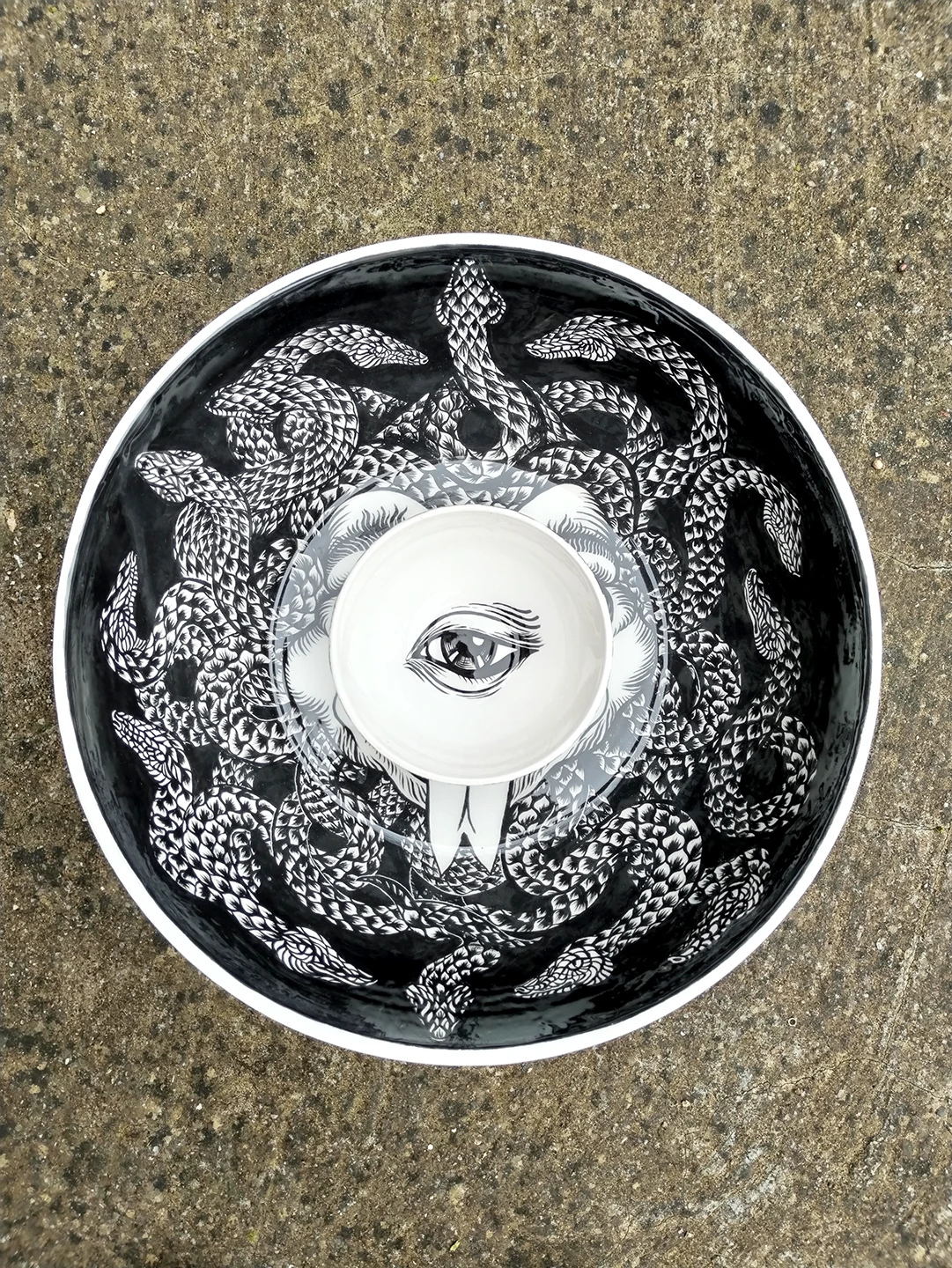 Foto che mostra le due ciotole del progetto "Medusa" posizionate l'una dentro l'altra su una superficie di pietra. La ciotola più piccola, decorata con il singolo occhio, è collocata al centro della ciotola grande, creando un gioco visivo in cui l'occhio sembra appartenere al volto della Gorgone sottostante, circondato dalla sua chioma di serpenti.