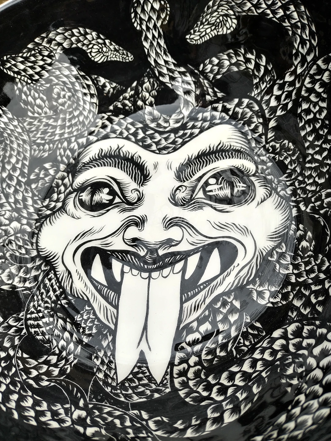 Dettaglio dell'opera in ceramica "Medusa" di Rosmunda che mostra il volto della Gorgone dipinto in bianco e nero sul fondo di una ciotola. L'illustrazione mette in risalto lo sguardo ipnotico, la bocca spalancata con denti aguzzi e lingua biforcuta sporgente, e la complessa trama di serpenti squamati che si intrecciano fittamente intorno al capo.