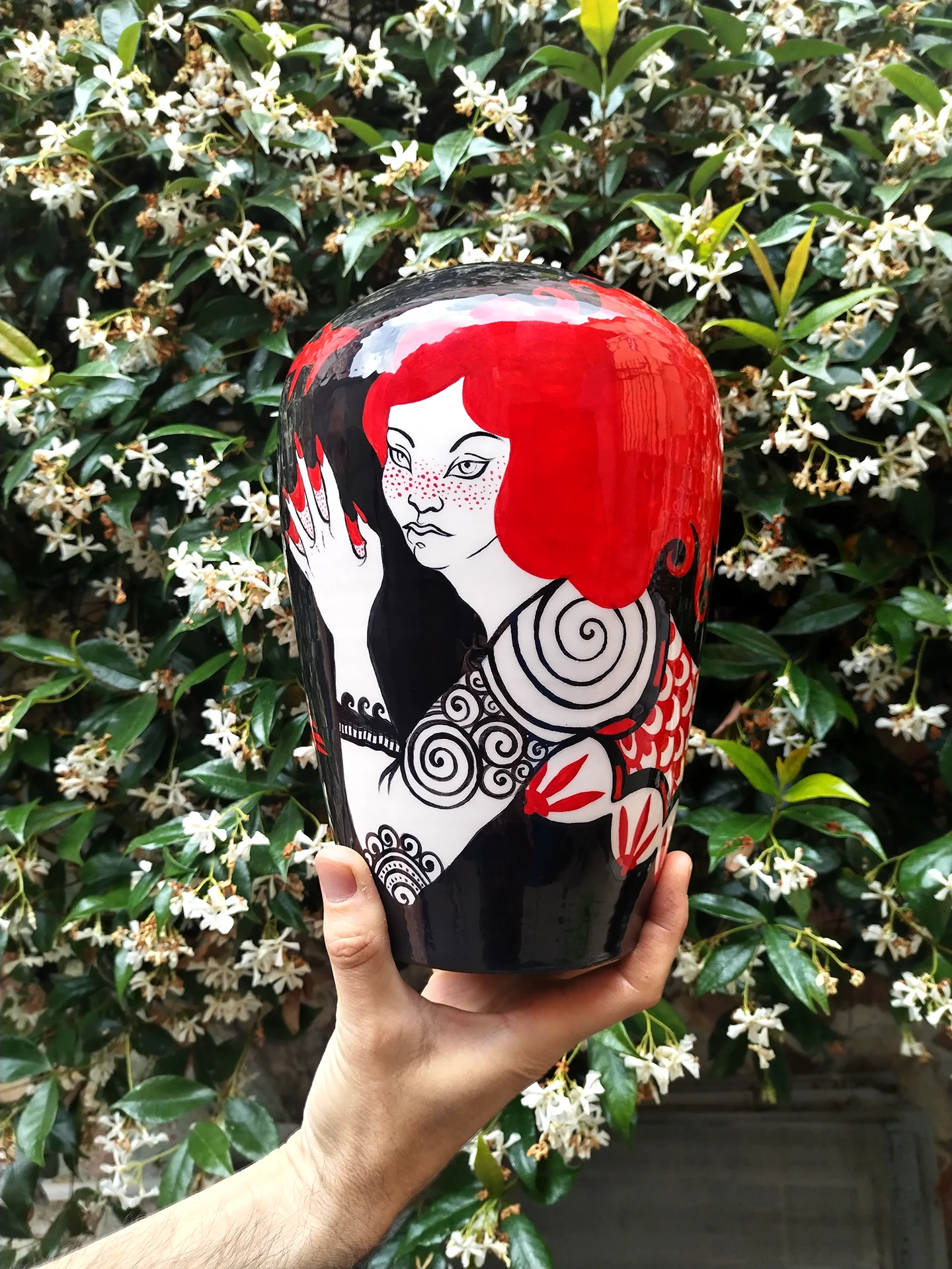 Foto che rappresenta una mano che mostra il vaso smaltato raffigurante una Sirena dai capelli rosso papavero e tatuaggi tribali sulle braccia, dita palmate seno coperto da fiori.