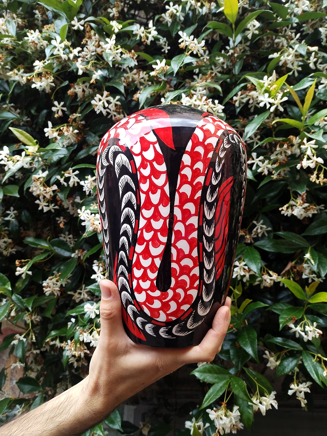 La coda squamata della Sirena scende sinuosa assecondando la curvatura del vaso. Il contrasto tra il rosso vibrante e il nero profondo viene esaltato dal candore della ceramica, creando un ritmo visivo armonioso che richiama la delicatezza dei fiori bianchi del gelsomino sullo sfondo.