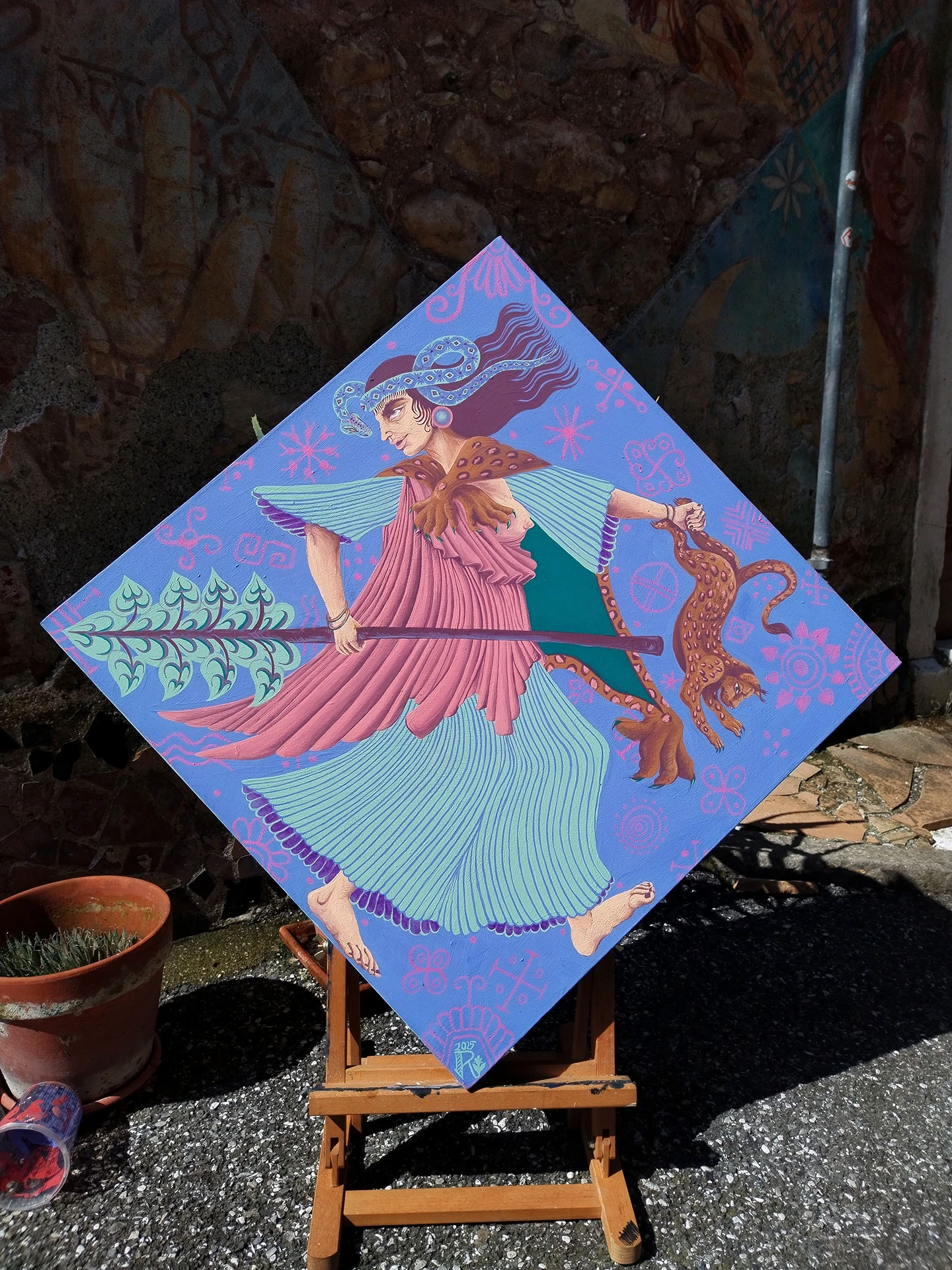 Foto dell'opera "Ekstasis" di Rosmunda. La tela romboidale è ancora sul cavalletto fuori dallo studio dell'artista: mostra la figura mitologica a mezzo busto su una tela romboidale azzurra posizionata su un cavalletto in legno all'aperto. La divinità indossa una veste rosa panneggiata che lascia scoperto un seno e una stola in pelle di leopardo, mentre impugna un bastone rituale. Il volto è incorniciato da un diadema a forma di serpente verde smeraldo con motivi a rombi, evidenziando il carattere sacro e selvaggio della composizione.