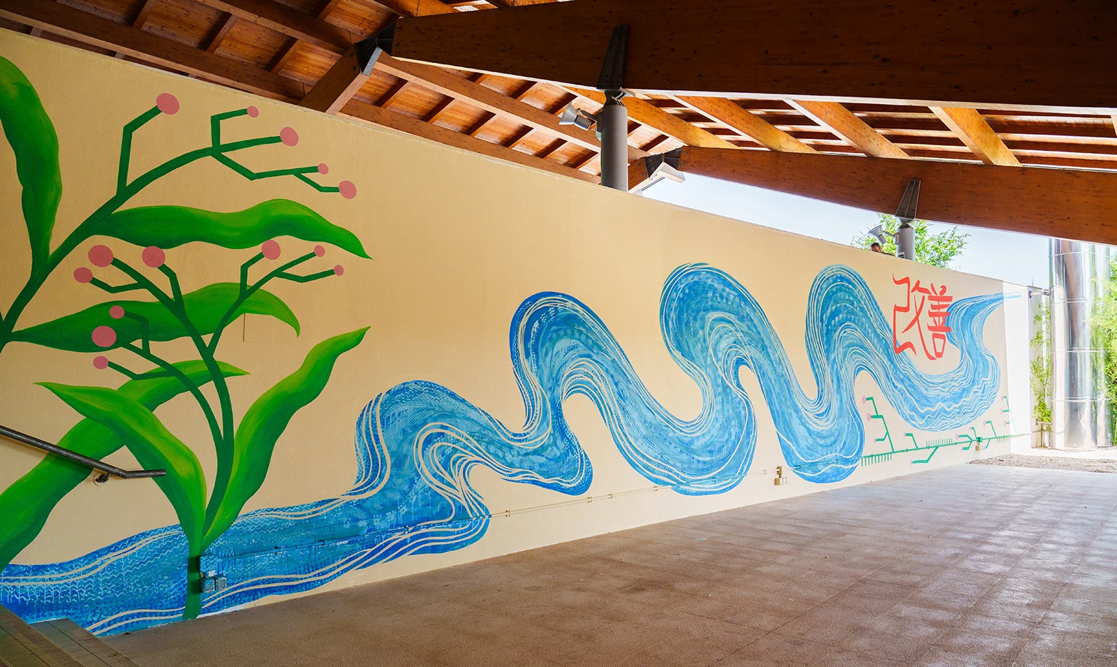 Veduta prospettica del murale realizzato per Toyota in un ambiente esterno coperto, caratterizzato da grandi piante verdi con frutti rosa che si integrano con onde blu stilizzate e iscrizioni calligrafiche rosse su una parete chiara.