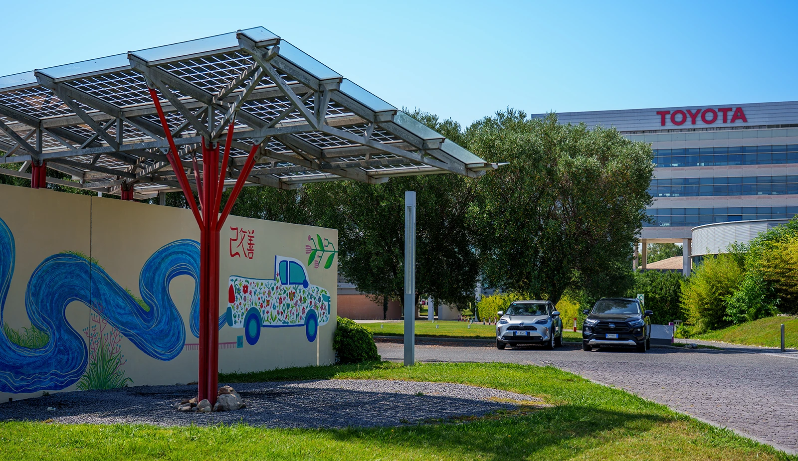 Veduta esterna del murale per Toyota in cui l'opera si integra con l'architettura circostante: una parete decorata con onde blu e l'illustrazione di un pick-up fiorito, situata accanto a una struttura metallica rossa che sostiene una tettoia solare.