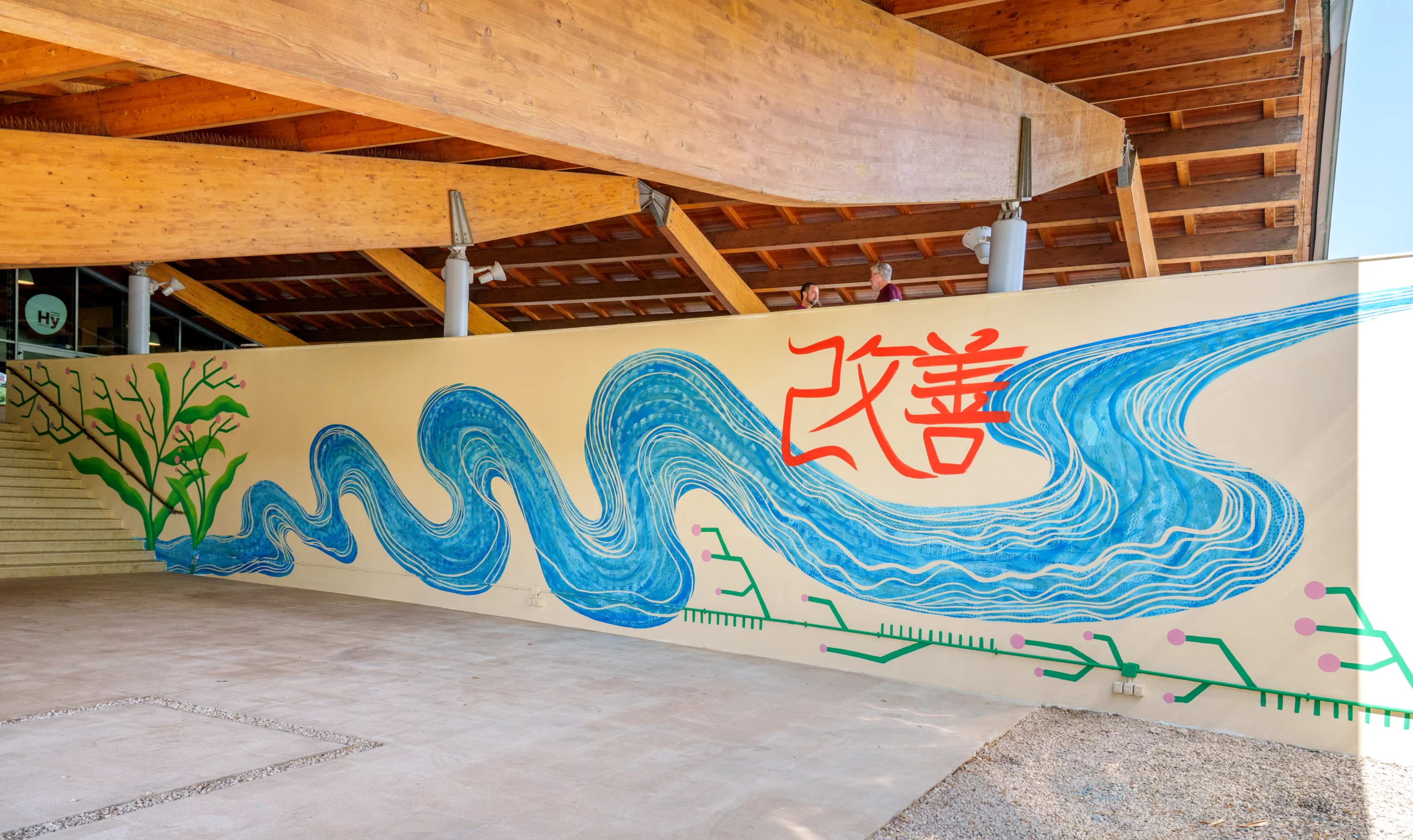 Veduta panoramica del murale realizzato per Toyota che mostra la progressione delle onde blu e degli elementi vegetali verdi lungo l'intera parete sotto la struttura in legno, con l'iscrizione calligrafica rossa centrale.