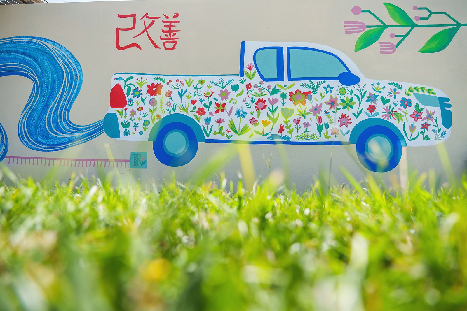 Illustrazione murale ravvicinata di un furgoncino pickup bianco e azzurro, decorato con una moltitudine di fiori variopinti e immerso in un prato d'erba verde, parte del murale per Toyota