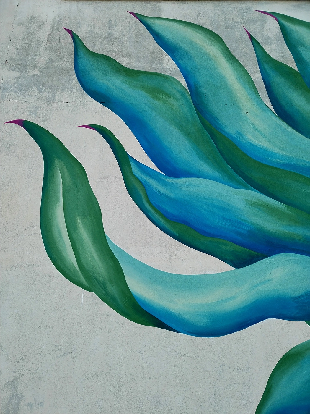 Dettaglio verticale del murale Agave di Rosmunda che mostra le lunghe foglie carnose e appuntite di una pianta di agave, dipinte con sfumature di verde, azzurro e blu che si intrecciano su uno sfondo chiaro.