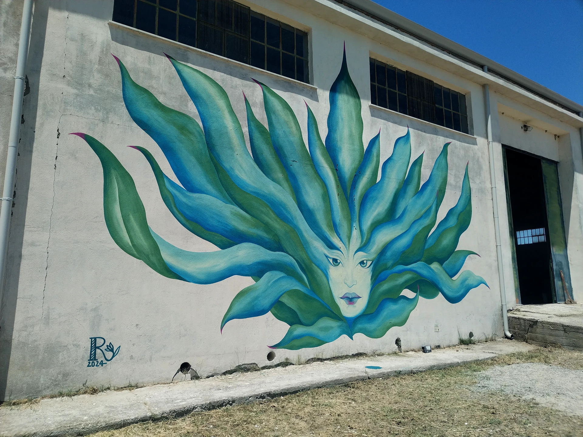 Dettaglio verticale del murale Agave di Rosmunda che ritrae un volto femminile stilizzato con grandi occhi azzurri, le cui fattezze si fondono con foglie carnose e appuntite nelle tonalità del verde e del blu, richiamando la forma di una pianta di agave.