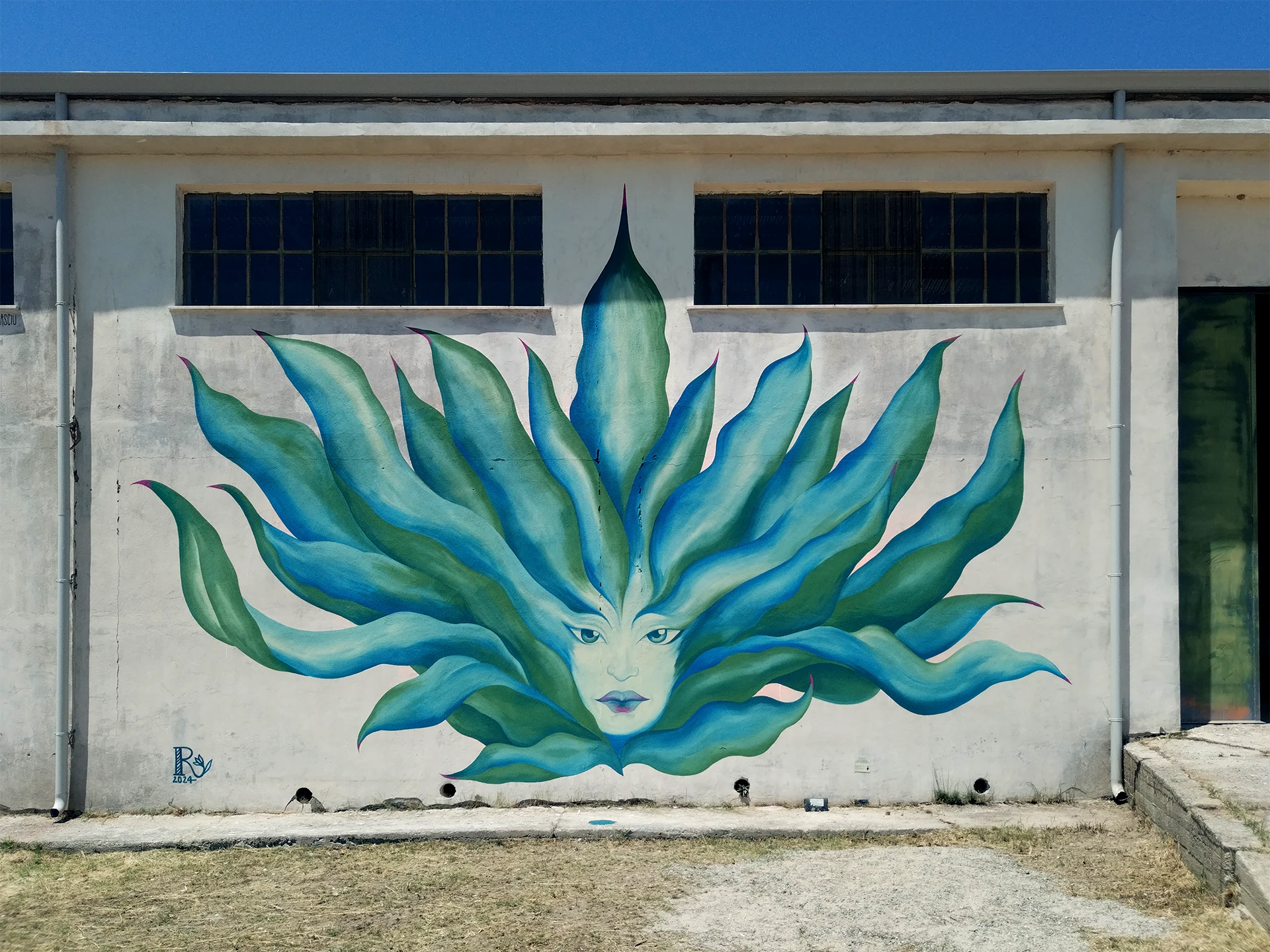 Veduta d'insieme del murale Agave di Rosmunda dipinto sulla facciata esterna di un edificio industriale, che raffigura un volto femminile le cui fattezze e i lunghi capelli si trasformano nelle foglie carnose e appuntite di una pianta di agave blu e verde.