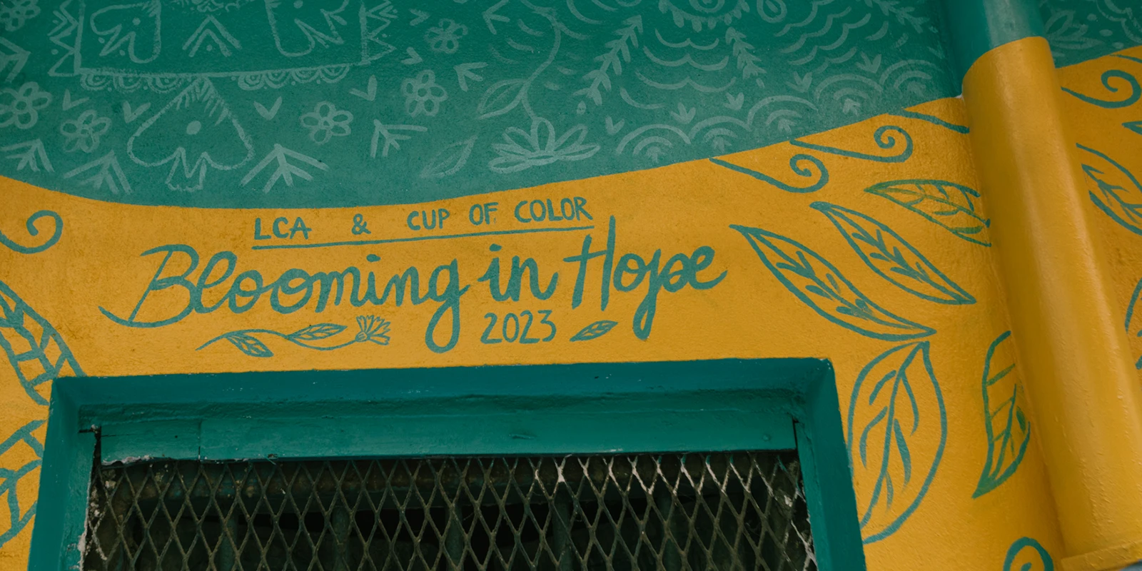 Dettaglio del murale "Blooming in Hope" di Rosmunda che mostra una porzione di muro giallo e azzurro con la scritta artistica "Blooming in Hope 2023" circondata da motivi floreali e fogliami stilizzati.