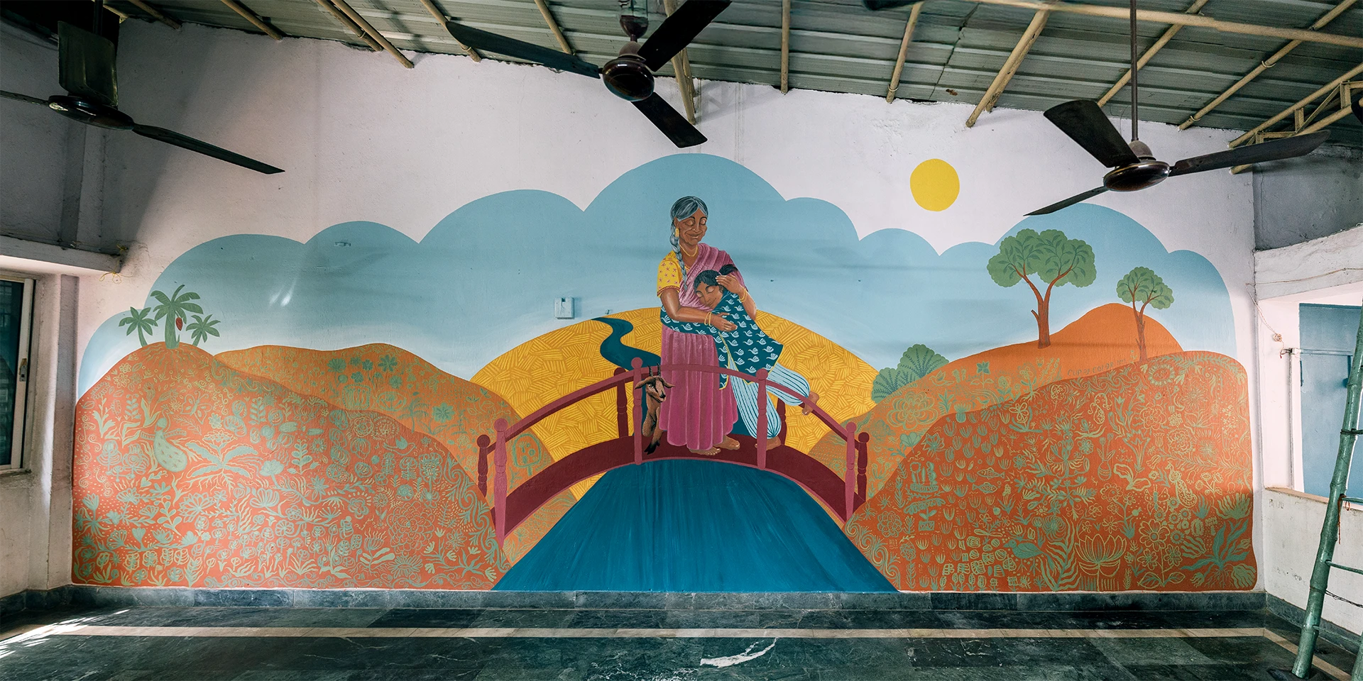 Veduta d'insieme del murale Embrace Your Inner Child di Rosmunda all'interno di una sala, che raffigura una donna in abiti tradizionali mentre abbraccia una bambina sopra un ponte rosso che attraversa un fiume blu, circondate da un paesaggio collinare dorato sotto un cielo azzurro con nuvole stilizzate.