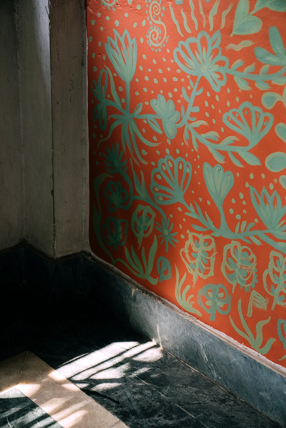Dettaglio verticale del murale Embrace Your Inner Child di Rosmunda che mostra una porzione di muro decorata con fitti motivi floreali stilizzati in verde acqua su un fondo arancione acceso, situata accanto a uno spigolo architettonico in ombra.