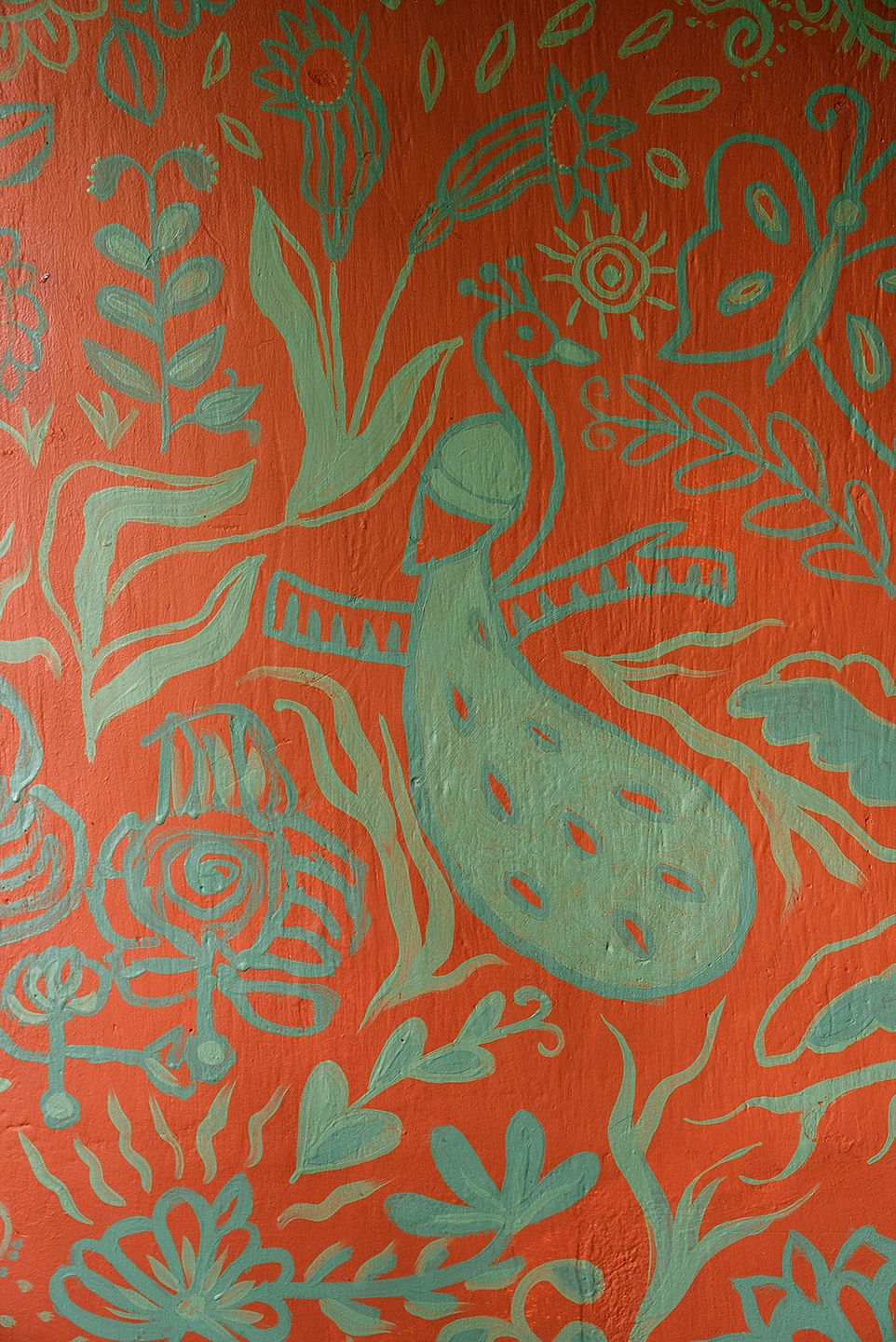 Dettaglio verticale del murale Embrace Your Inner Child di Rosmunda che raffigura un pavone verde acqua stilizzato tra motivi floreali e decorativi della stessa tonalità, dipinti su un fondo arancione vivace.