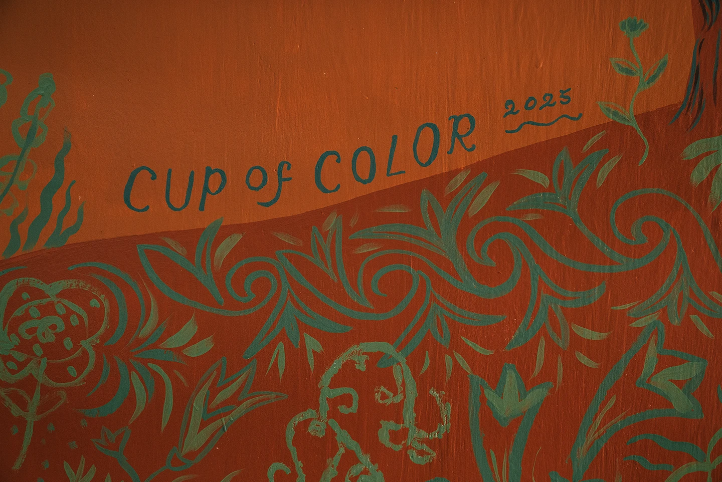 Dettaglio del murale Embrace Your Inner Child di Rosmunda che mostra la firma della ONG "CUP OF COLOR" e l'anno "2025" scritti in azzurro su una collina arancione, decorata con complessi motivi floreali stilizzati in verde acqua.