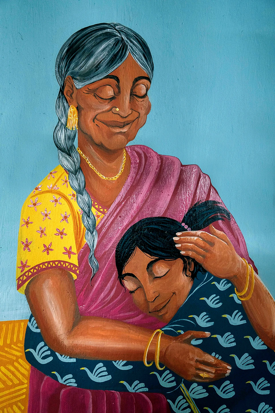Dettaglio verticale del murale Embrace Your Inner Child di Rosmunda che ritrae una donna anziana mentre accarezza la testa di una bambina che si appoggia al suo petto; la donna ha capelli grigi raccolti e indossa un sari rosa e giallo, mentre la bambina ha una veste azzurra decorata con piccoli uccelli bianchi.