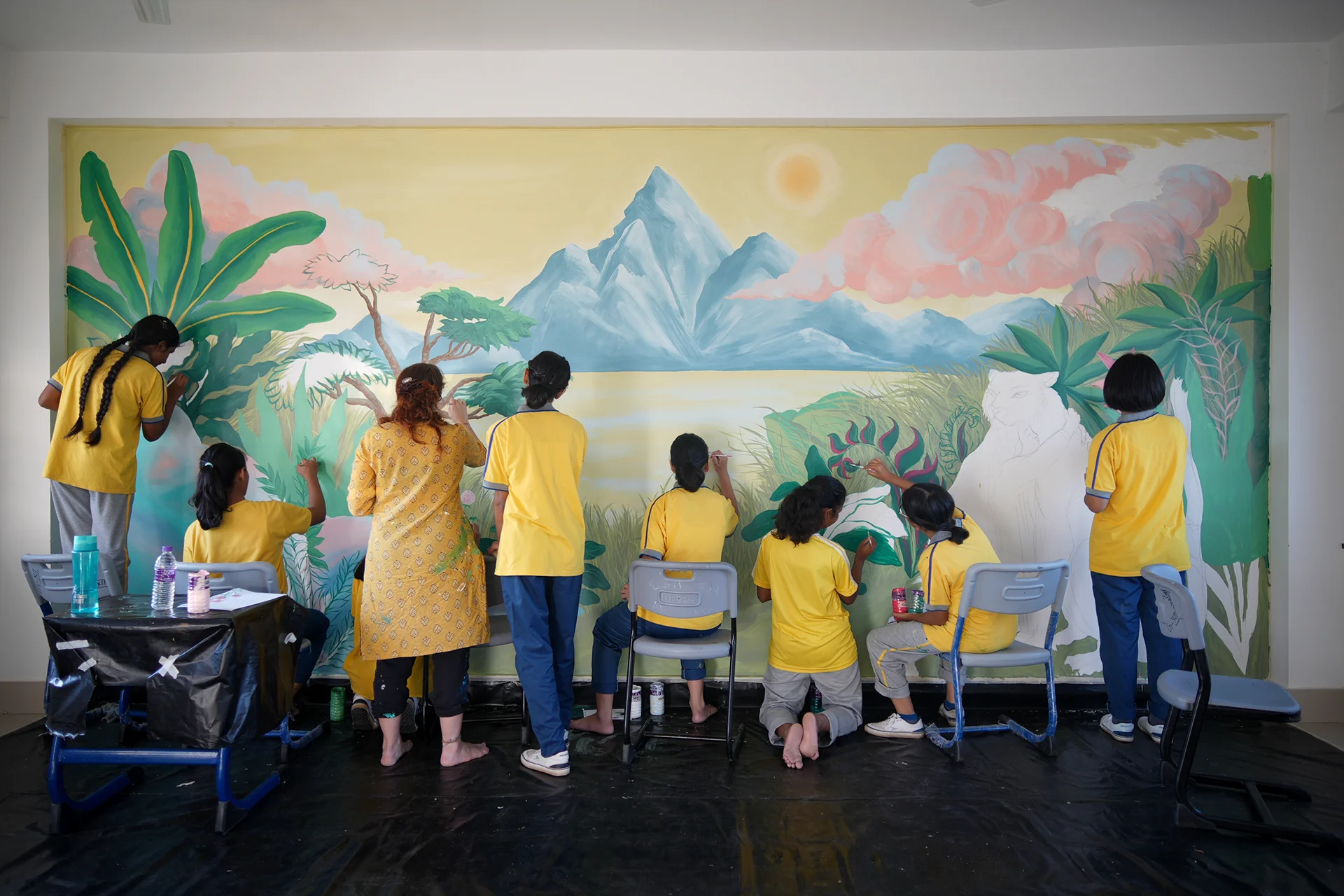 Fotografia che ritrae un gruppo di studentesse indiane in divisa gialla mentre dipingono un grande murale su una parete interna, collaborando alla creazione di un paesaggio naturale con montagne blu, un cielo dorato e una fitta giungla ricca di dettagli vegetali.