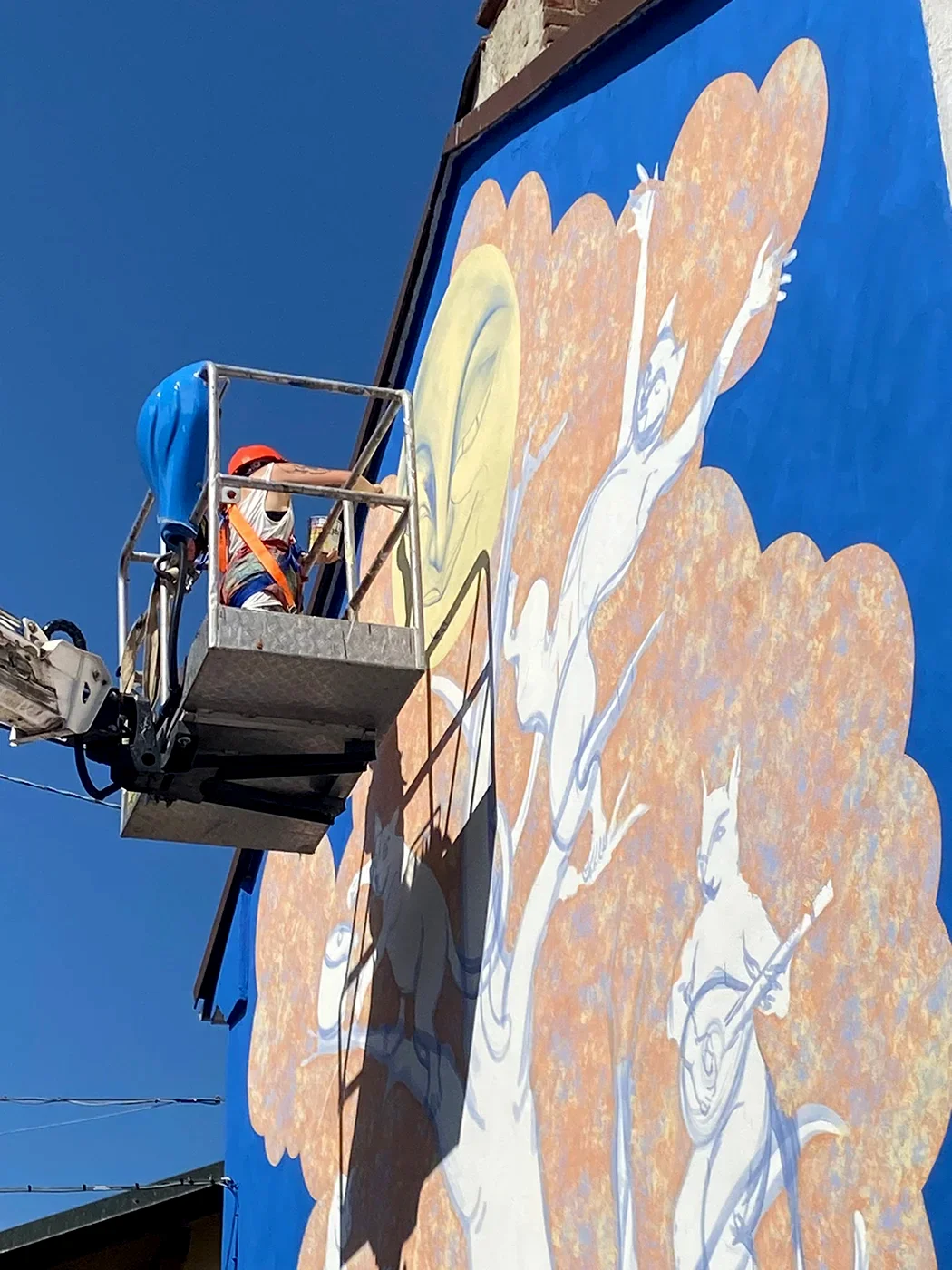 Dettaglio del murale "Il Segreto di Villa Chioso" che mostra un gatto bianco antropomorfo che suona uno strumento a corde, dipinto su un albero stilizzato accanto a una figura umana che si protende verso l'alto, con l'artista al lavoro su una piattaforma elevatrice.