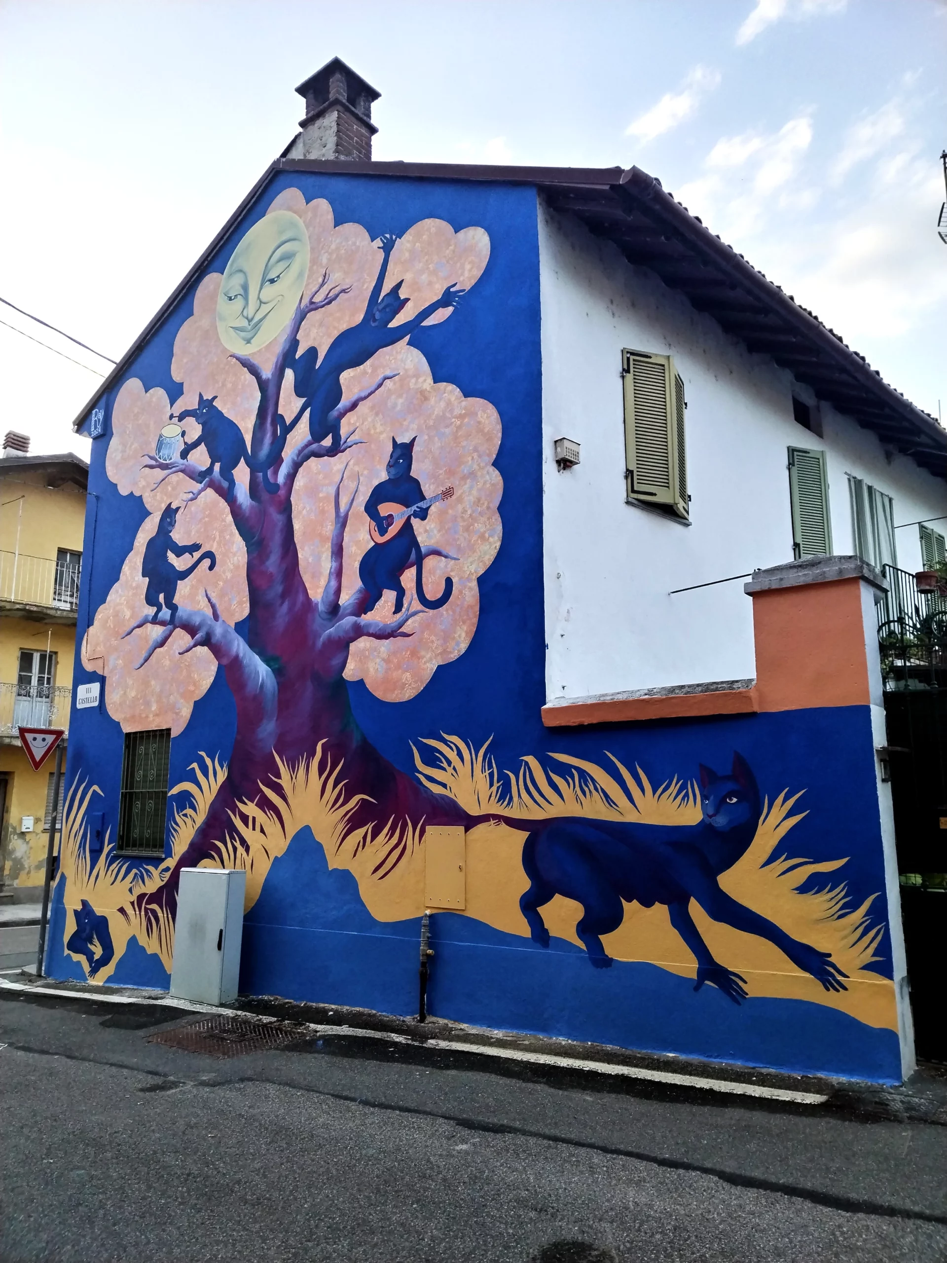 Veduta d'insieme del murale "Il Segreto di Villa Chioso" sulla facciata di un edificio, che raffigura un grande albero stilizzato con gatti blu antropomorfi e un volto di luna piena antropomorfo sulla sommità, sovrastante una silhouette di gatto blu che corre in primo piano su uno sfondo giallo e blu.