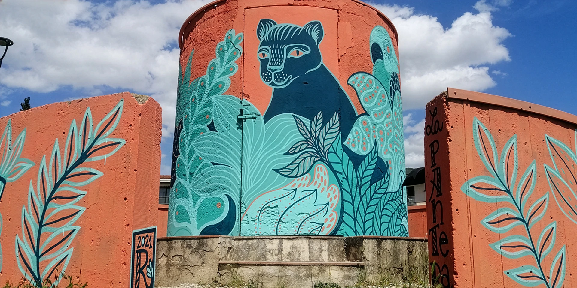 Veduta panoramica del murale "La Pantera" di Rosmunda, realizzato su una struttura cilindrica e muri perimetrali arancioni, che raffigura una pantera blu circondata da fitta vegetazione stilizzata in varie tonalità di azzurro e verde sotto un cielo parzialmente nuvoloso.