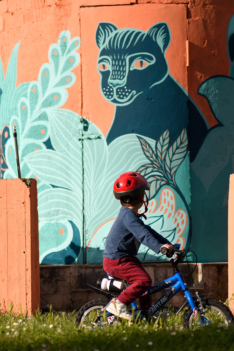 Dettaglio del murale "La Pantera" di Rosmunda che mostra un bambino con casco rosso mentre pedala su una bicicletta blu davanti a una parete arancione decorata con una pantera stilizzata di colore blu e foglie azzurre.