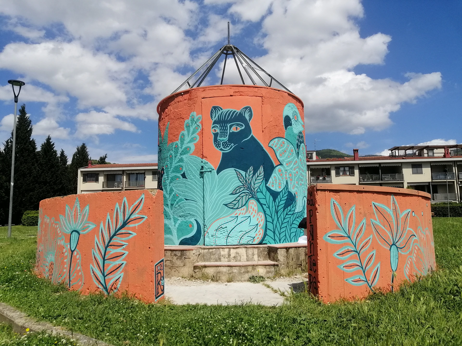 Veduta panoramica del murale "La Pantera" di Rosmunda, realizzato su una struttura cilindrica e muri perimetrali arancioni, che raffigura una pantera blu circondata da fitta vegetazione stilizzata in varie tonalità di azzurro e verde sotto un cielo parzialmente nuvoloso.