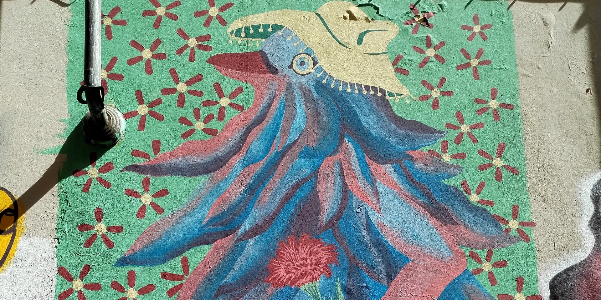 Veduta orizzontale del murale 'Piccione Viaggiatore' di Rosmunda: un uccello antropomorfo piumato con piumaggio blu e rosso, che indossa un cappello di paglia decorato, circondato da piccoli fiori rossi su un fondo verde menta.