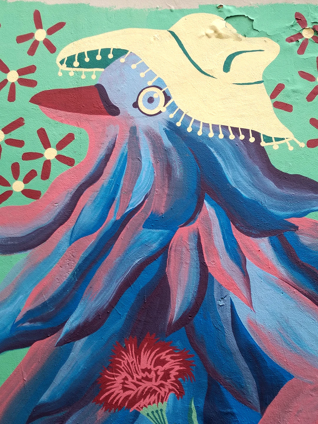 Dettaglio del murale 'Piccione Viaggiatore' di Rosmunda che ritrae la testa dell'uccello antropomorfo con piumaggio blu e rosso, mentre indossa un cappello di paglia decorato, su uno sfondo verde menta con fiori stilizzati.