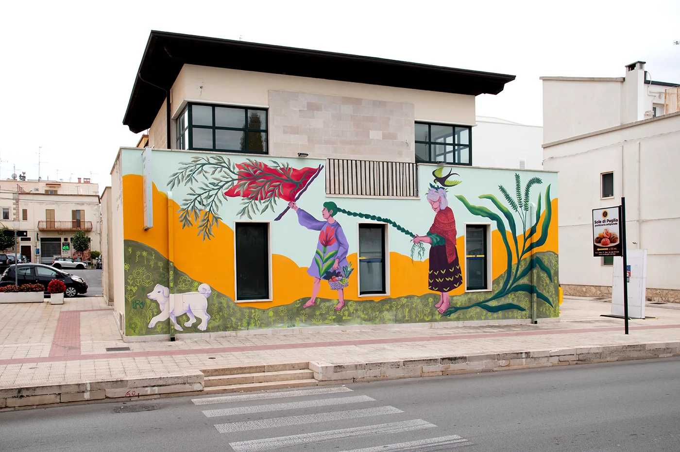 Murale del progetto "Radici Erranti" di Rosmunda che raffigura, su una facciata urbana, un cane bianco, una figura che tiene un grande elemento vegetale rosso e una donna anziana con capelli viola, integrati in un paesaggio stilizzato con colline gialle e foglie verdi.
