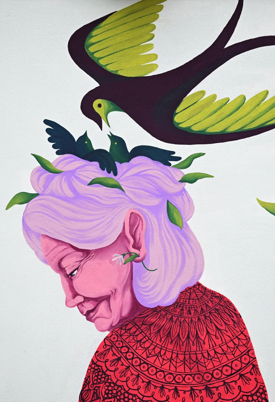 Illustrazione artistica del progetto "Radici Erranti" di Rosmunda che ritrae il profilo di una donna anziana con capelli viola raccolti, decorati da foglie e piccoli uccelli neri, mentre un grande uccello variopinto vola sopra la sua testa; la figura indossa una veste rossa con fitti motivi ornamentali neri su sfondo bianco.