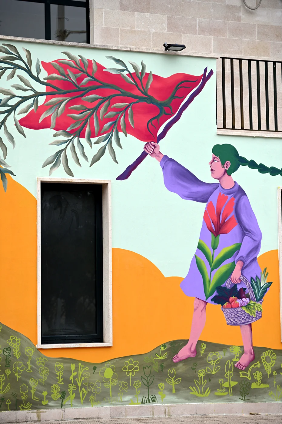 Dettaglio del murale "Radici Erranti" che raffigura una giovane donna di profilo con una lunga treccia verde, vestita con un abito lilla decorato da un grande fiore rosso, mentre solleva un ramo con foglie rosse e tiene un cesto di prodotti della terra, su uno sfondo a colline arancioni e gialle.