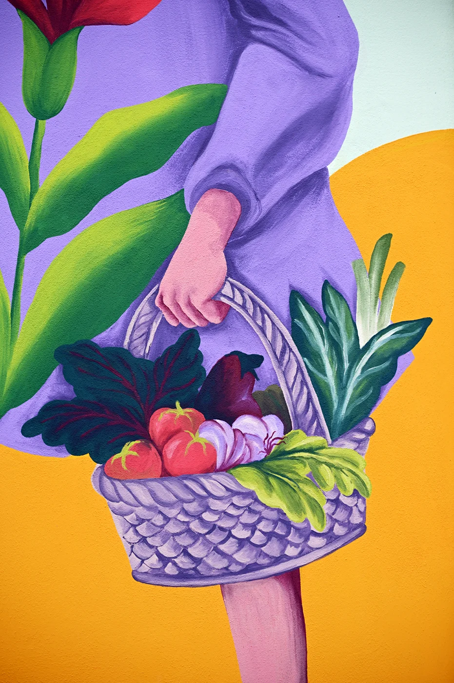 Dettaglio del murale "Radici Erranti" di Rosmunda che inquadra in primo piano un cesto intrecciato ricolmo di ortaggi freschi, tra cui pomodori e verdure a foglia, sorretto da una figura che indossa una veste lilla su uno sfondo a colline gialle e arancioni.