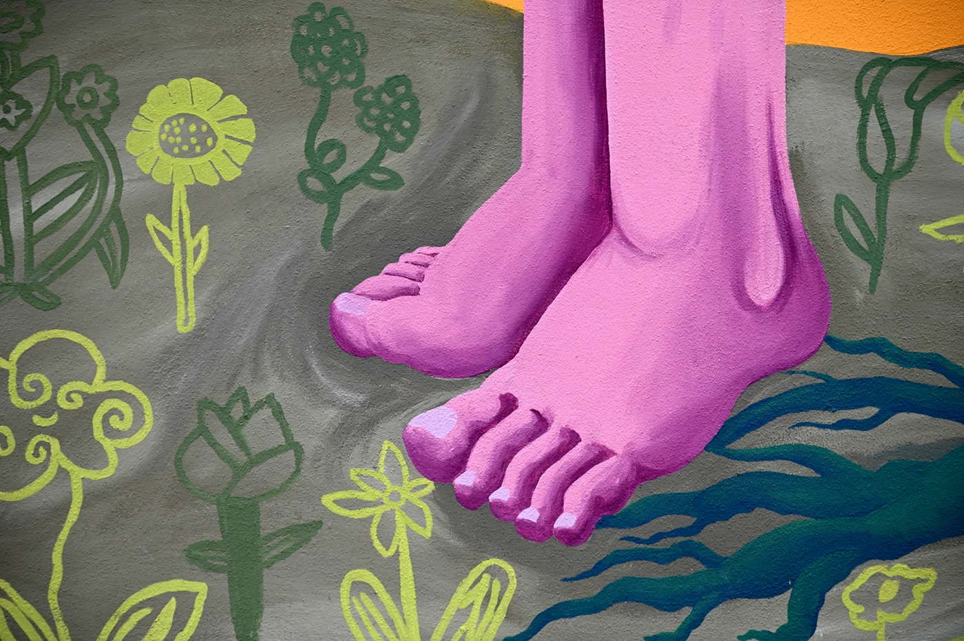 Dettaglio del murale "Radici Erranti" di Rosmunda che raffigura un paio di piedi nudi di colore rosa/lilla che camminano su un terreno grigio decorato con fiori stilizzati gialli e verdi.