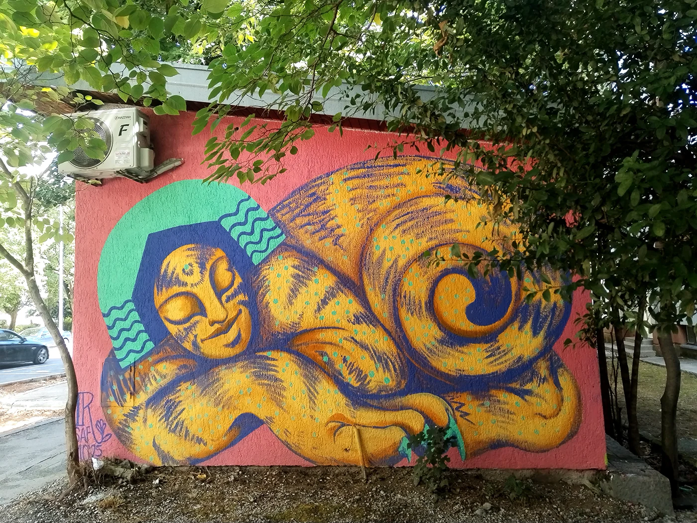 Veduta d'insieme del murale 'Sphinx' di Rosmunda: una monumentale figura mitologica distesa che occupa l'intera parete esterna, con il volto dorato e il corpo decorato da pattern gialli e blu che contrastano con lo sfondo rosa acceso dell'edificio.
