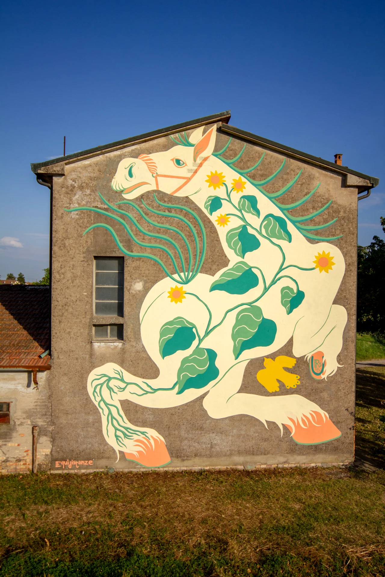 Veduta prospettica centrale del murale "Tormento, Cavallo Contento" di Rosmunda che mostra un grande cavallo bianco stilizzato con ali verdi e decorazioni floreali gialle, dipinto sulla facciata laterale di un edificio rurale.
